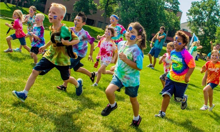 Children in tie-died shirts and face paints racing each other across a sunny field.