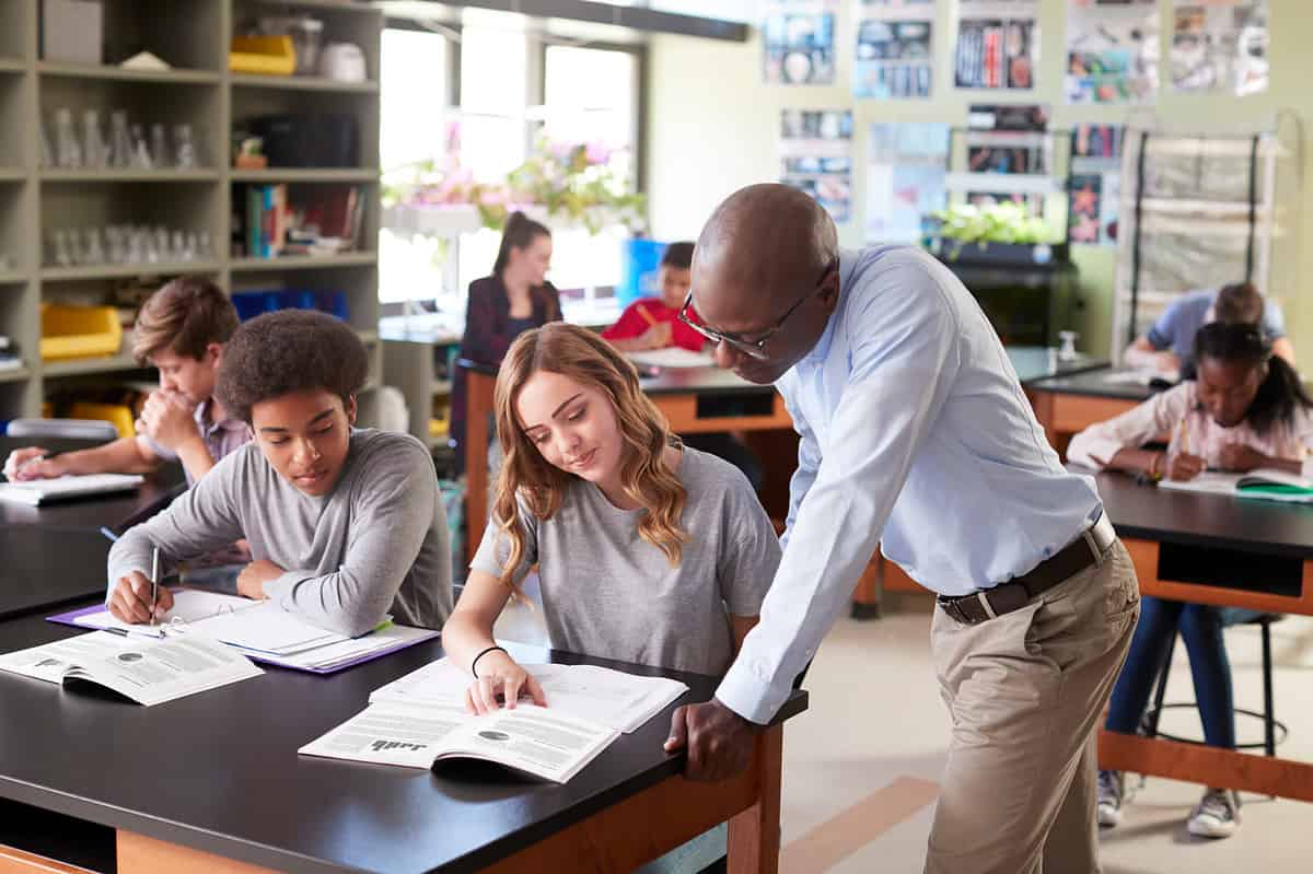 A high school teacher leaning in to explain a topic to a pupil in class.