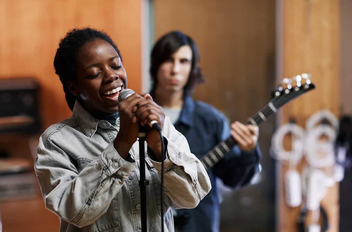Waist up portrait of young black woman singing to microphone while rehearsing with band in professional recording studio