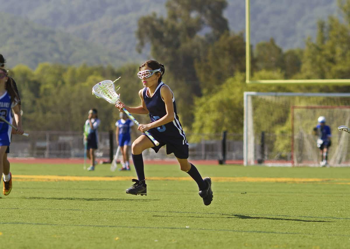 A young girl runs on a lacrosse field. She is carrying her lacrosse stick. The photo captures her in mid air.