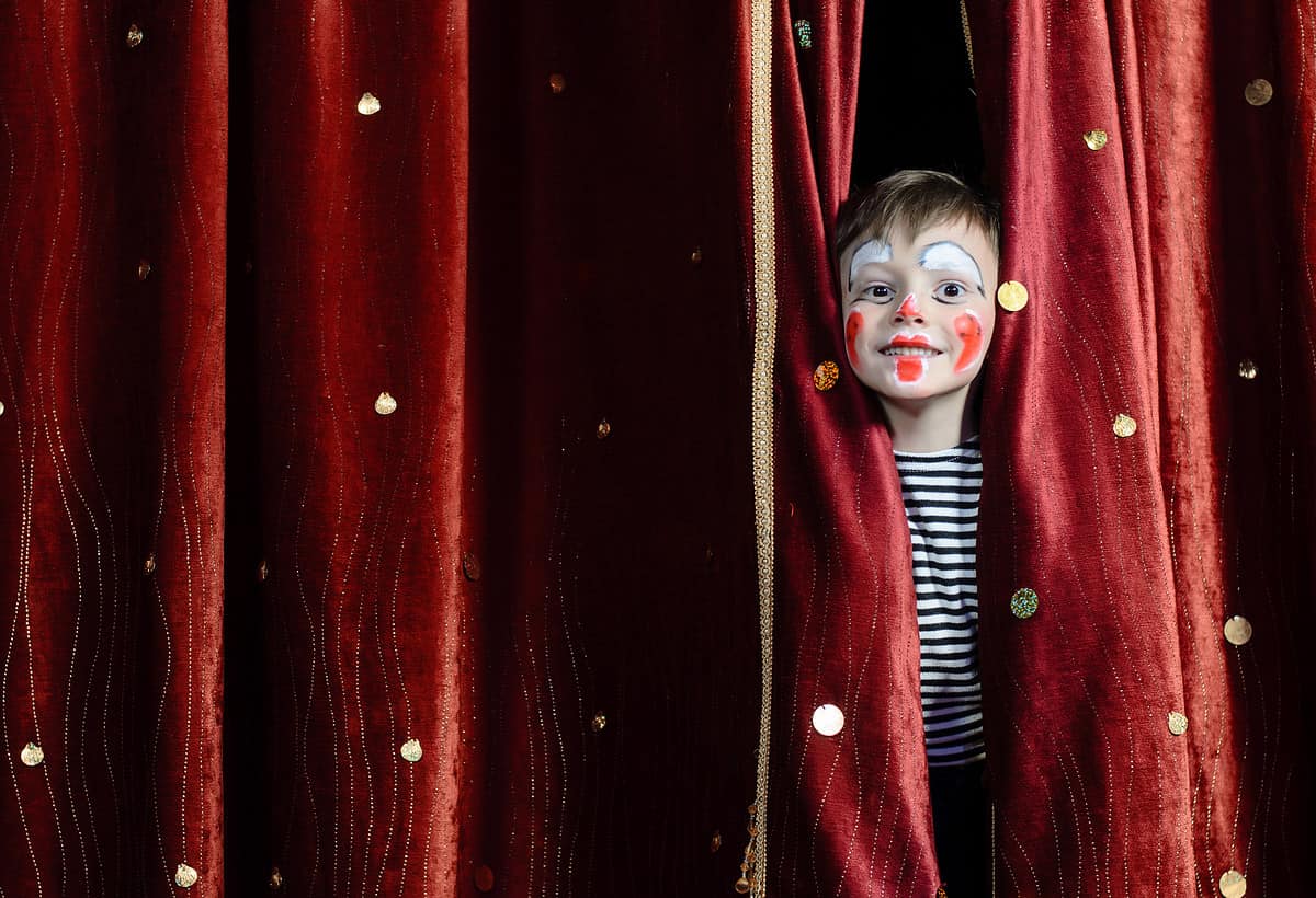 Young Boy Wearing Clown Make Up Peering Out Through Opening in Red Stage Curtains