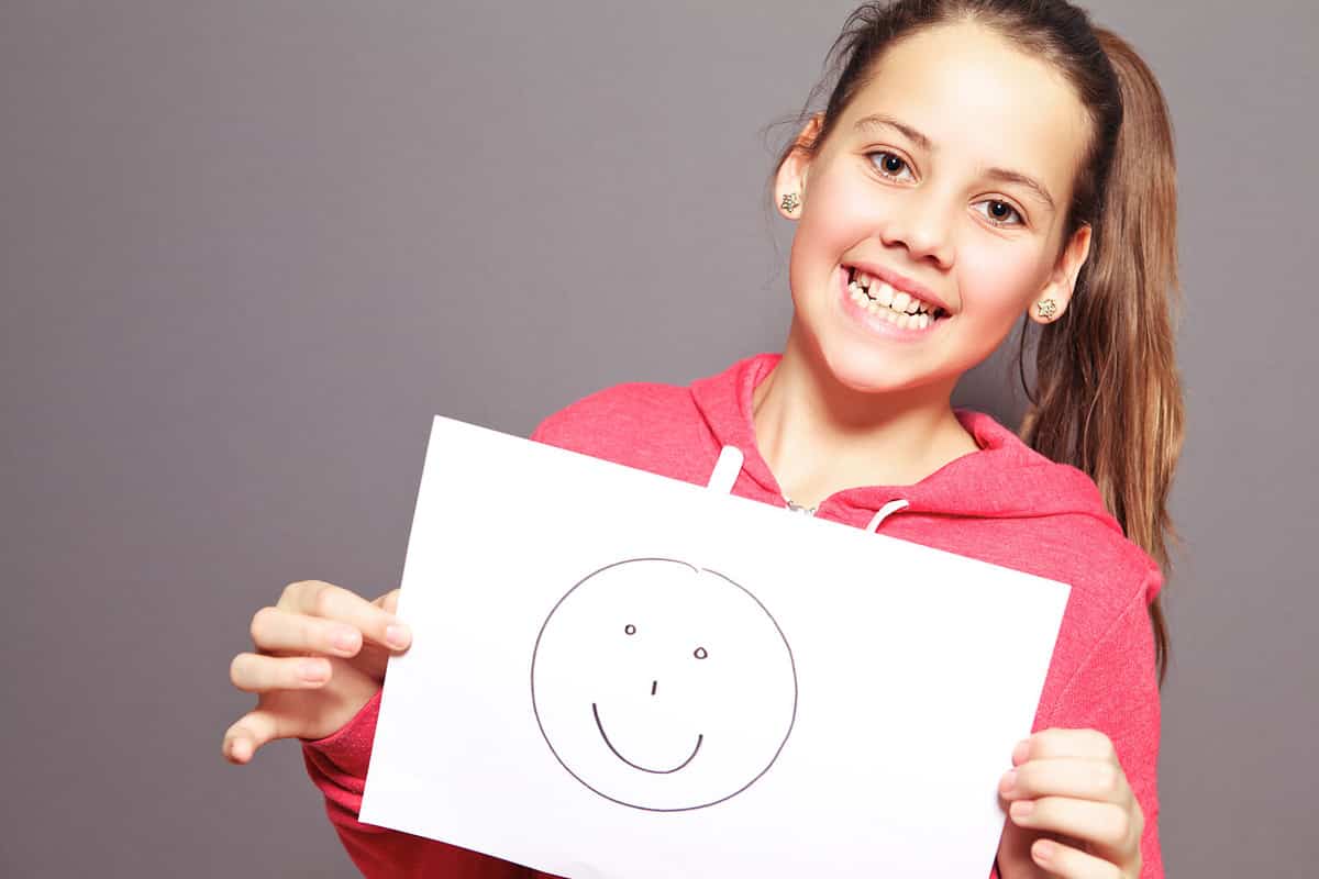 Happy attractive young girl with a lovely toothy smile holding a smiley drawn on a sheet of white paper, half body studio portrait on grey
