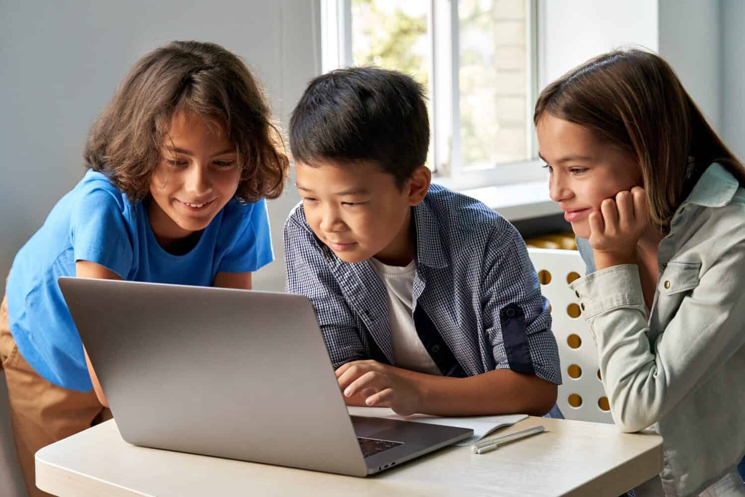 Diverse happy school kids using laptop computer together in classroom. Multicultural children junior students classmates learning online elementary education program class gathered at desk.