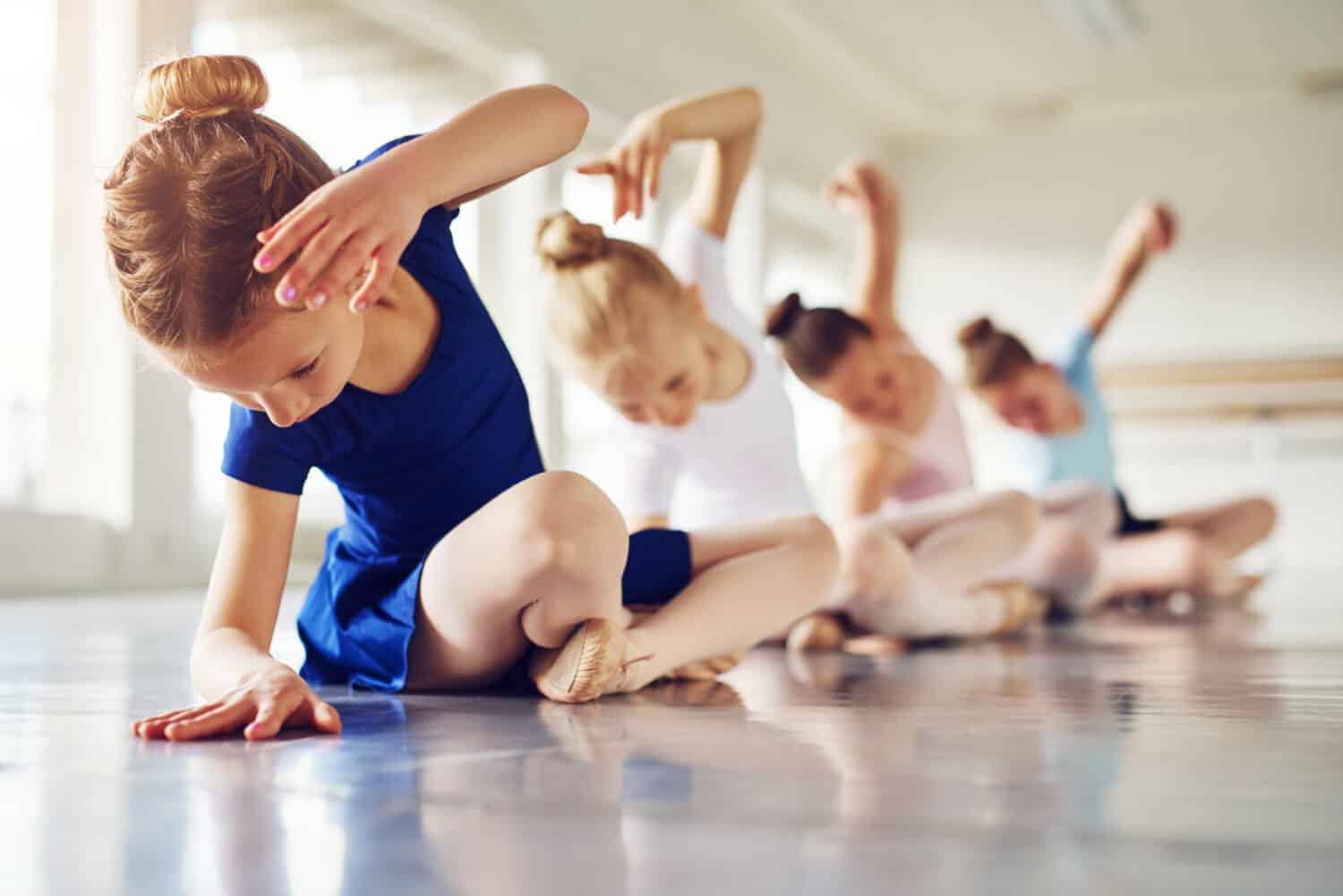 Little ballerinas doing exercises and bending sitting on floor in ballet class.