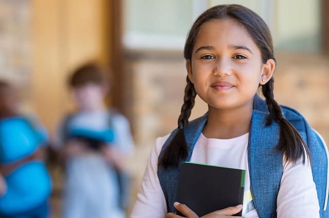 Smiling student girl wearing school backpack and holding exercise book. Portrait of happy asian young girl outside the primary school. Closeup face of smiling hispanic schoolgirl looking at camera.