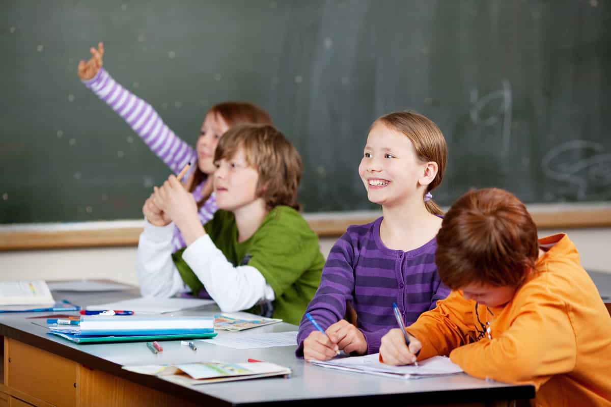 Smiling friendly little girl in class looking at the front of the classroom while one of her friends waves her hand in the air to answer a question