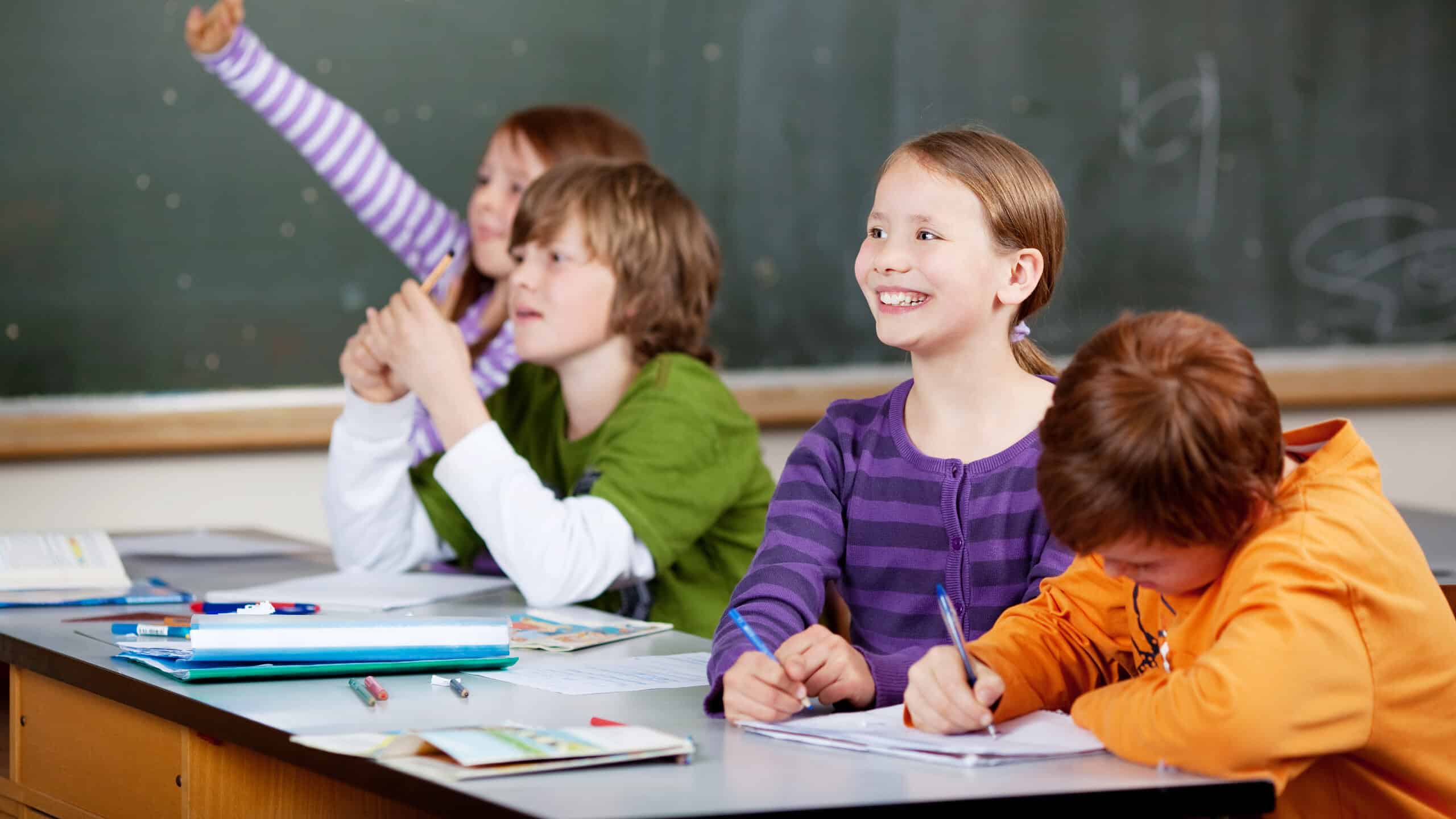 Smiling friendly little girl in class looking at the front of the classroom while one of her friends waves her hand in the air to answer a question