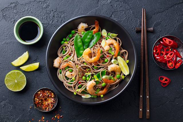 Stir fry noodles with vegetables and shrimps in black bowl. Slate background. Top view