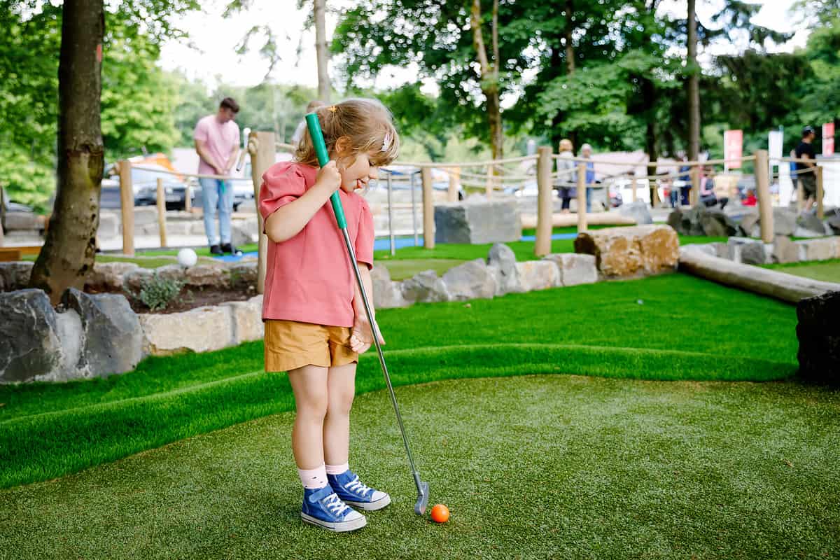 Cute preschool girl playing mini golf with family. Happy toddler child having fun with outdoor activity. Summer sport for children and adults, outdoors. Family vacations or resort