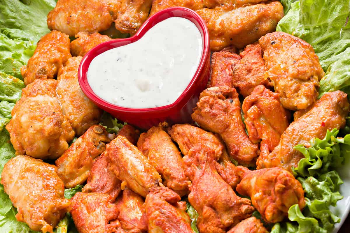 "A high angle close up of a plate full of Buffalo wings with a red, heart shaped bowl full of blue cheese dipping sauce."