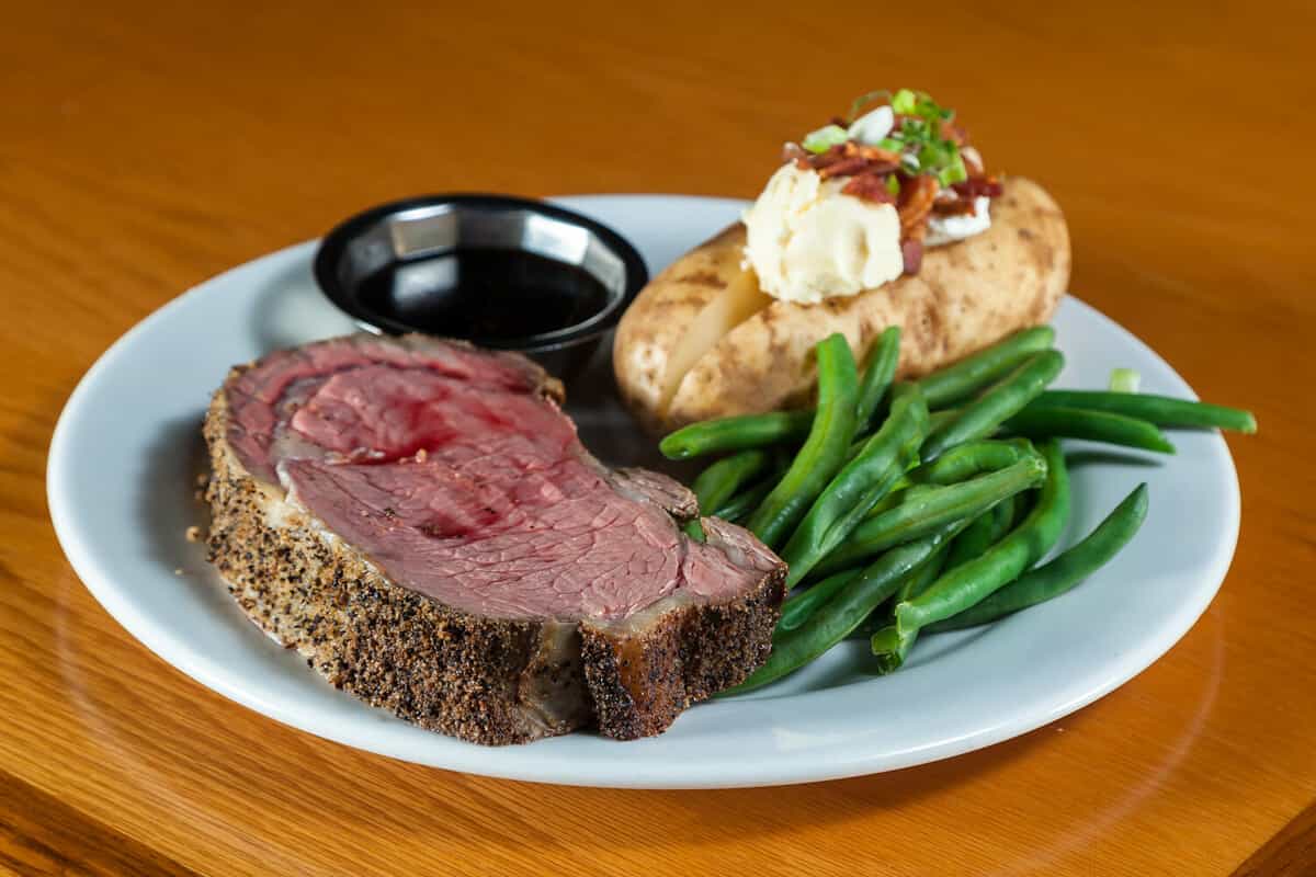 prime rib meal served on a plate with a loaded potato and green beans with a side of a jus