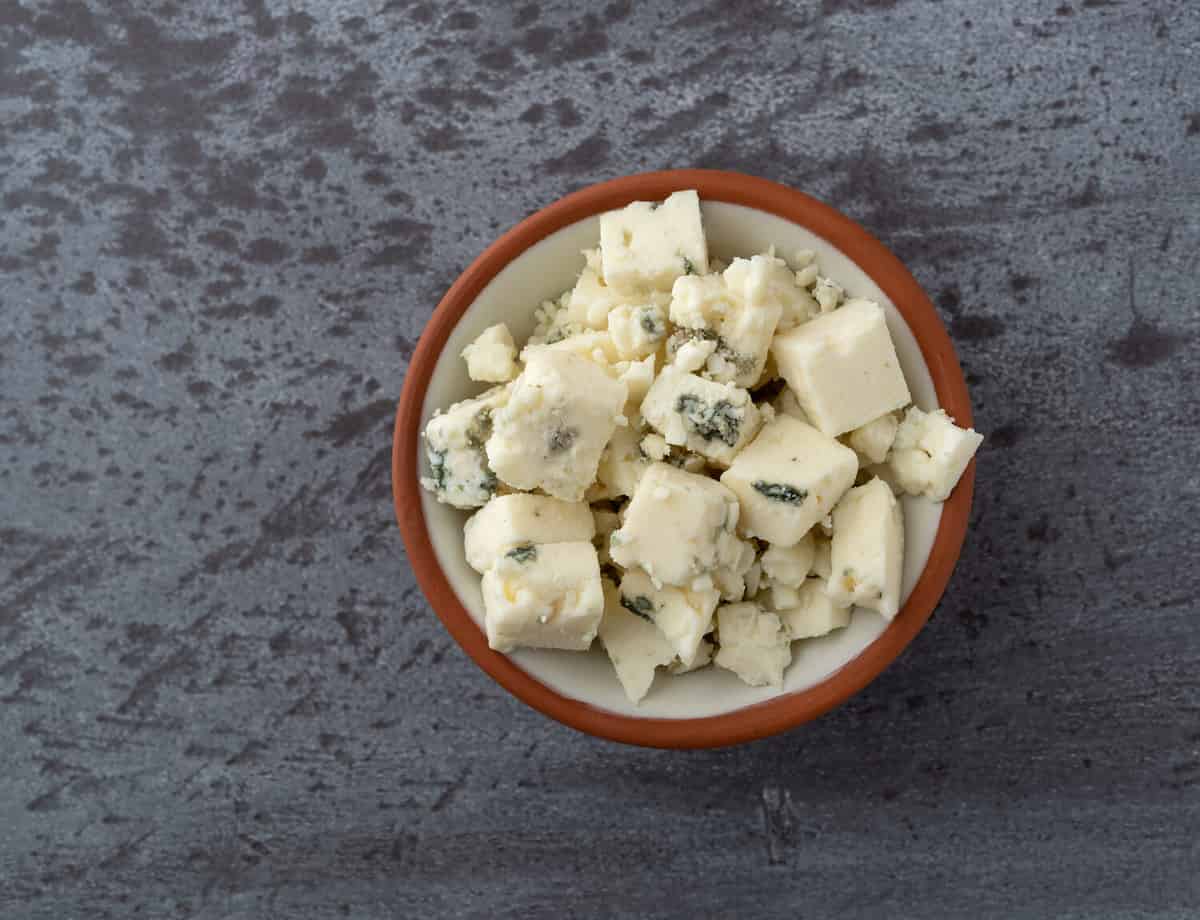 Overhead view of a portion of crumbled blue cheese in a small bowl atop a gray background illuminated with natural lighting.