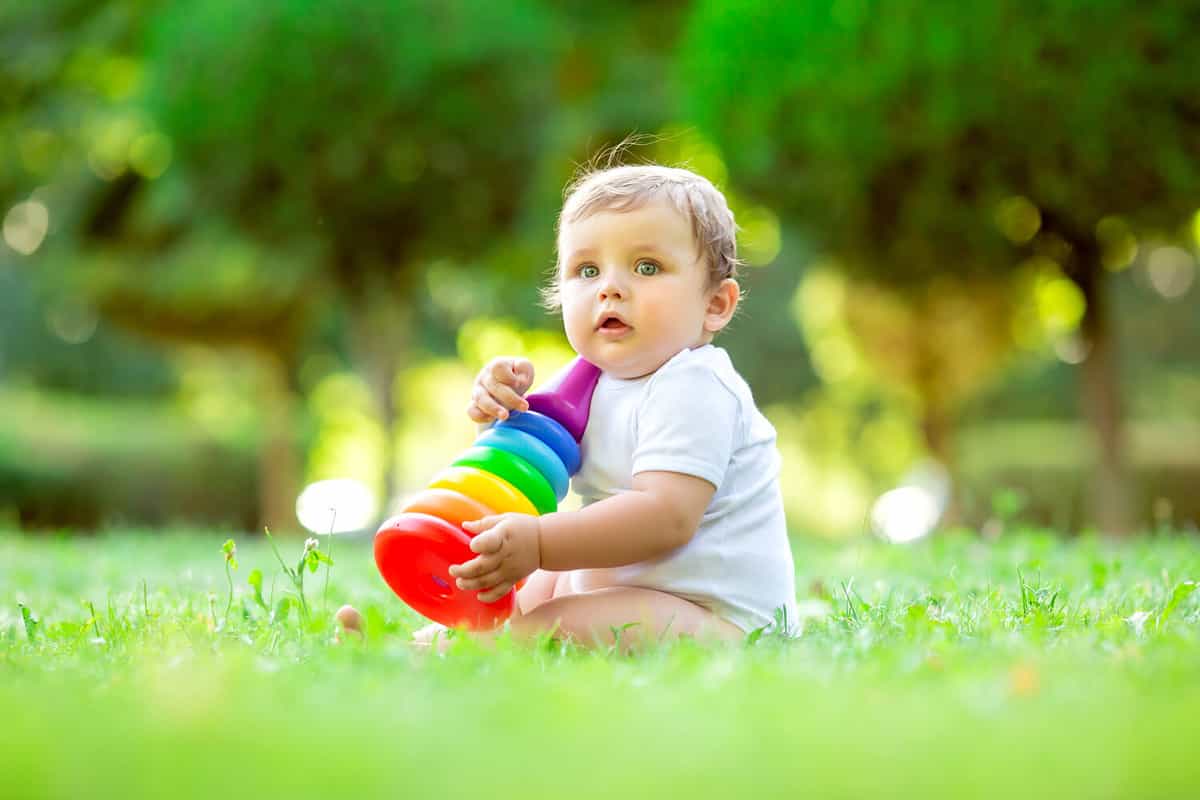 Cute toddler boy in white bodysuit sitting on the green grass in summer plays with a dress multi-colored pyramid. The child plays with the developing toy. Breaking up a child up to a year
