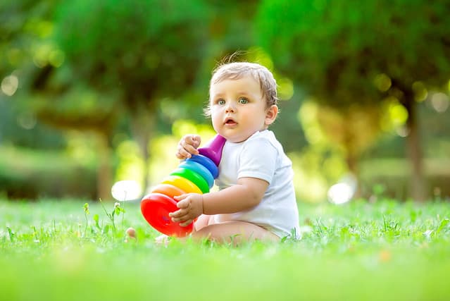 Cute toddler boy in white bodysuit sitting on the green grass in summer plays with a dress multi-colored pyramid. The child plays with the developing toy. Breaking up a child up to a year