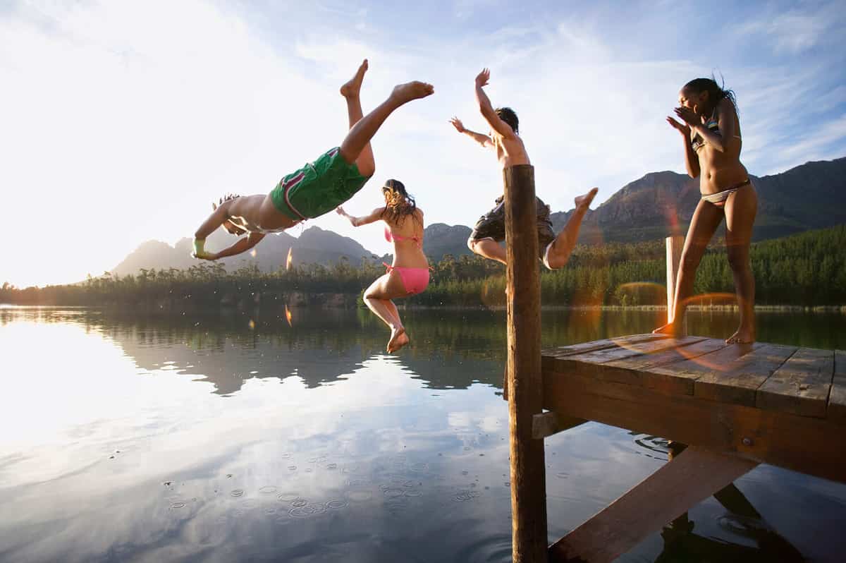 Horizontal shot of three adults jumping off a jetty into a lake as a girl in bikini cheers them with copy space.