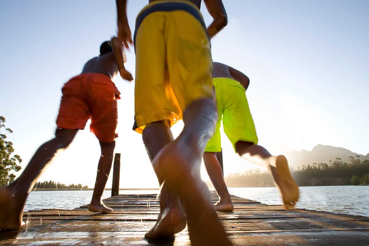 Young boys in swimming shorts running on a wooden path for a dive in the lake.