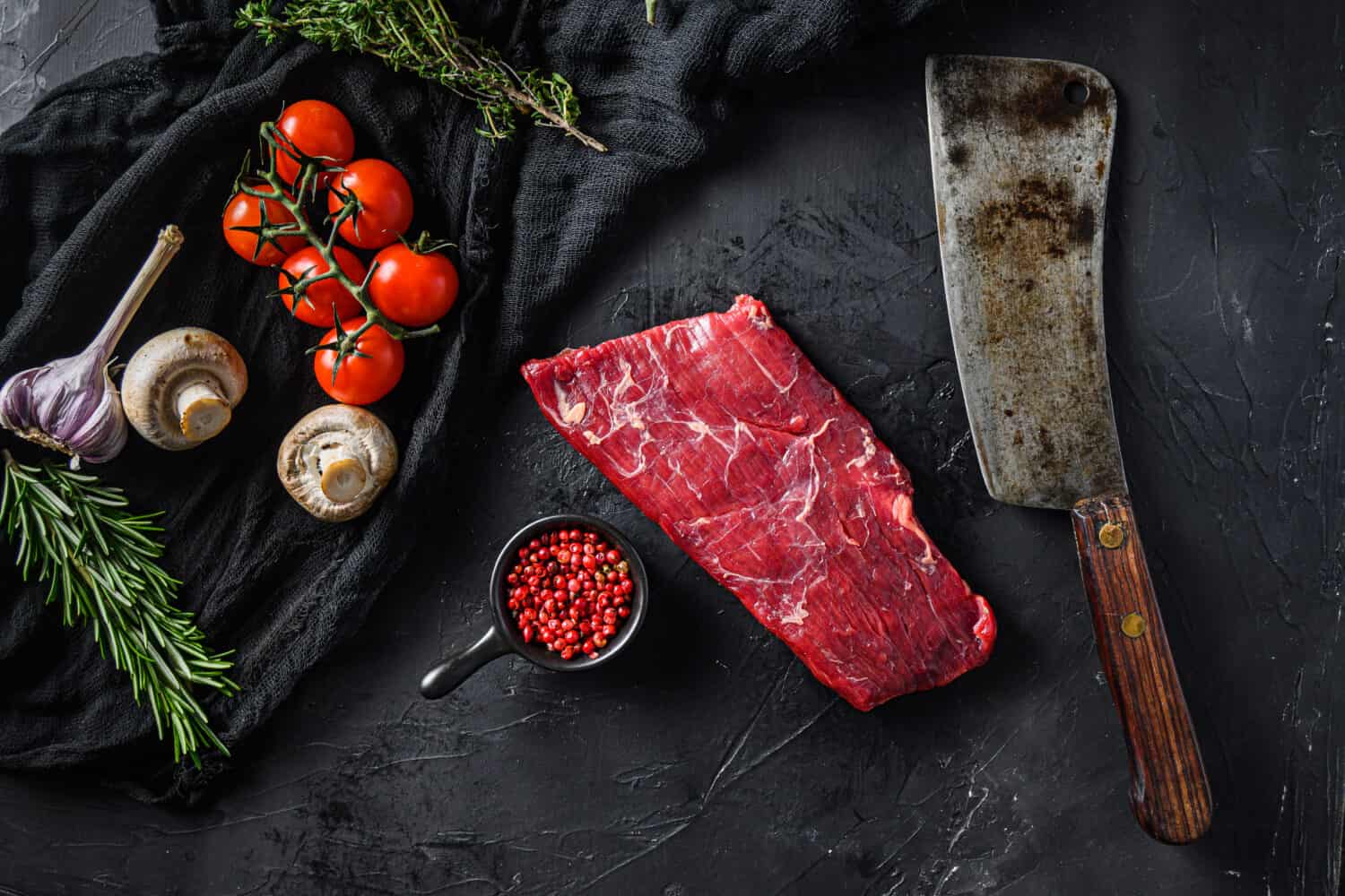 Raw, flap or flank, also known Bavette steak near butcher knife with pink pepper and rosemary. Black background. Top view.