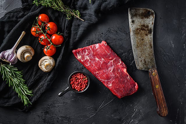 Raw, flap or flank, also known Bavette steak near butcher knife with pink pepper and rosemary. Black background. Top view.