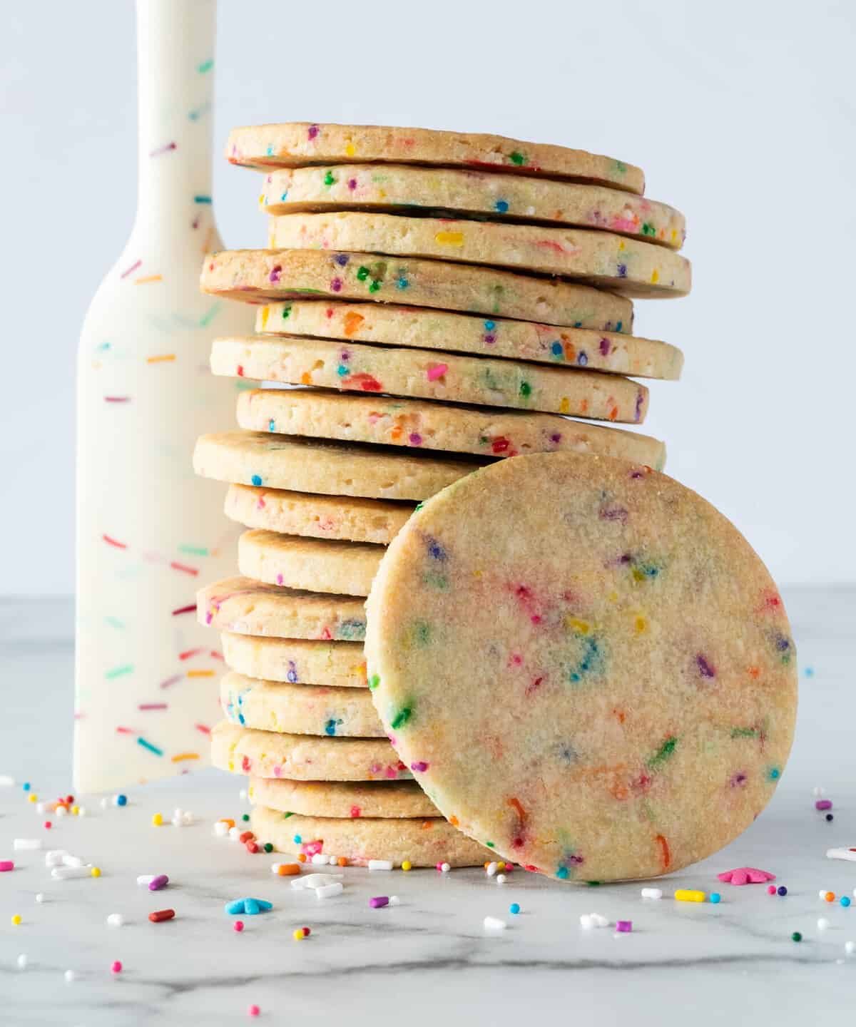 A stack of sprinkle sugar cookies with a sprinkle designed rubber spatula standing beside and sprinkles scattered.