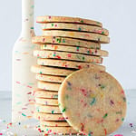 A stack of sprinkle sugar cookies with a sprinkle designed rubber spatula standing beside and sprinkles scattered.