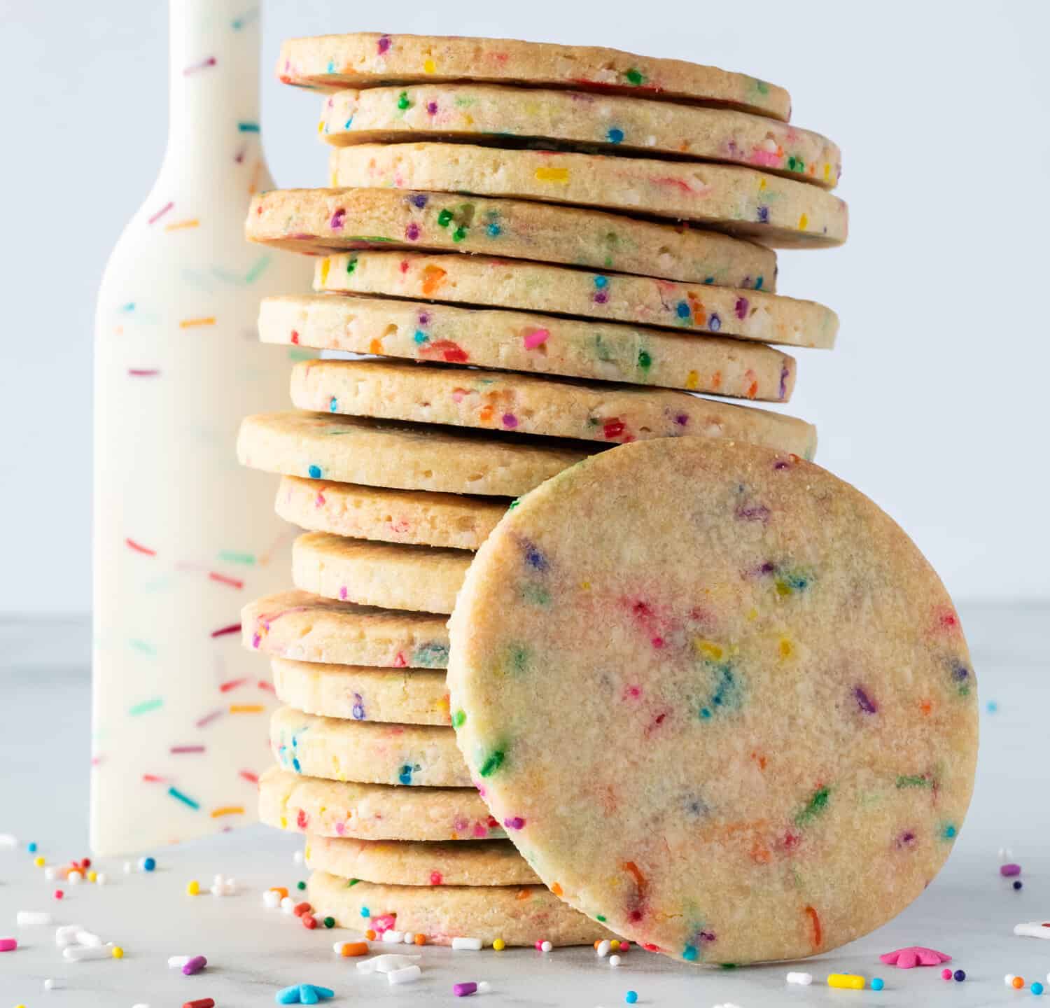 A stack of sprinkle sugar cookies with a sprinkle designed rubber spatula standing beside and sprinkles scattered.