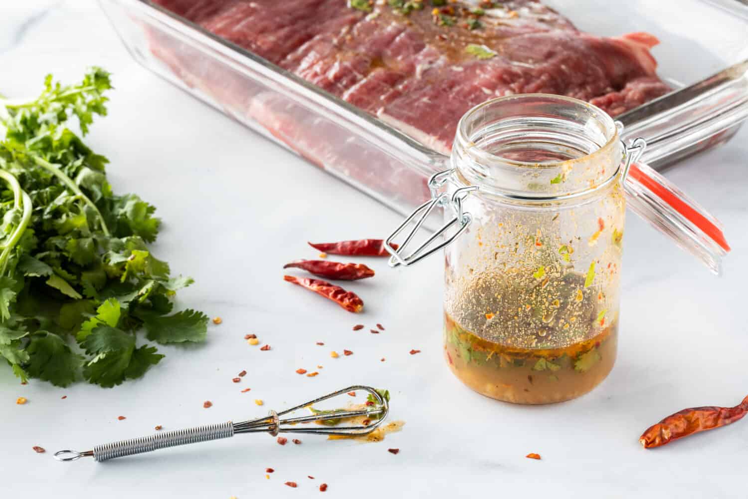 Close up of a jar of lime and cilantro marinade with cilantro herb on the side, and a flank of raw beef in behind.