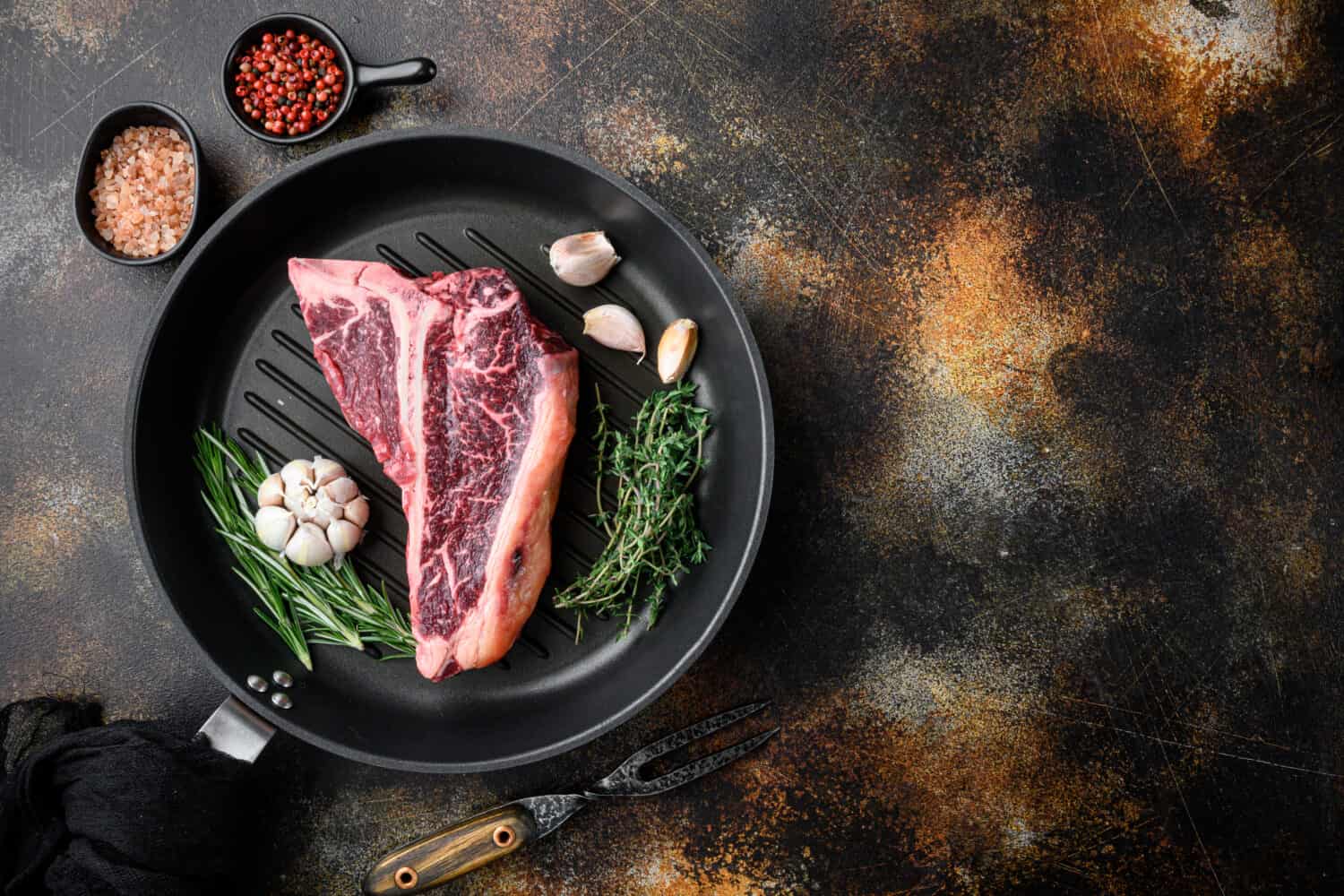 Big raw porterhouse steak with spices and ingredients, on frying cast iron pan, on old dark rustic background, top view flat lay, with copy space for text