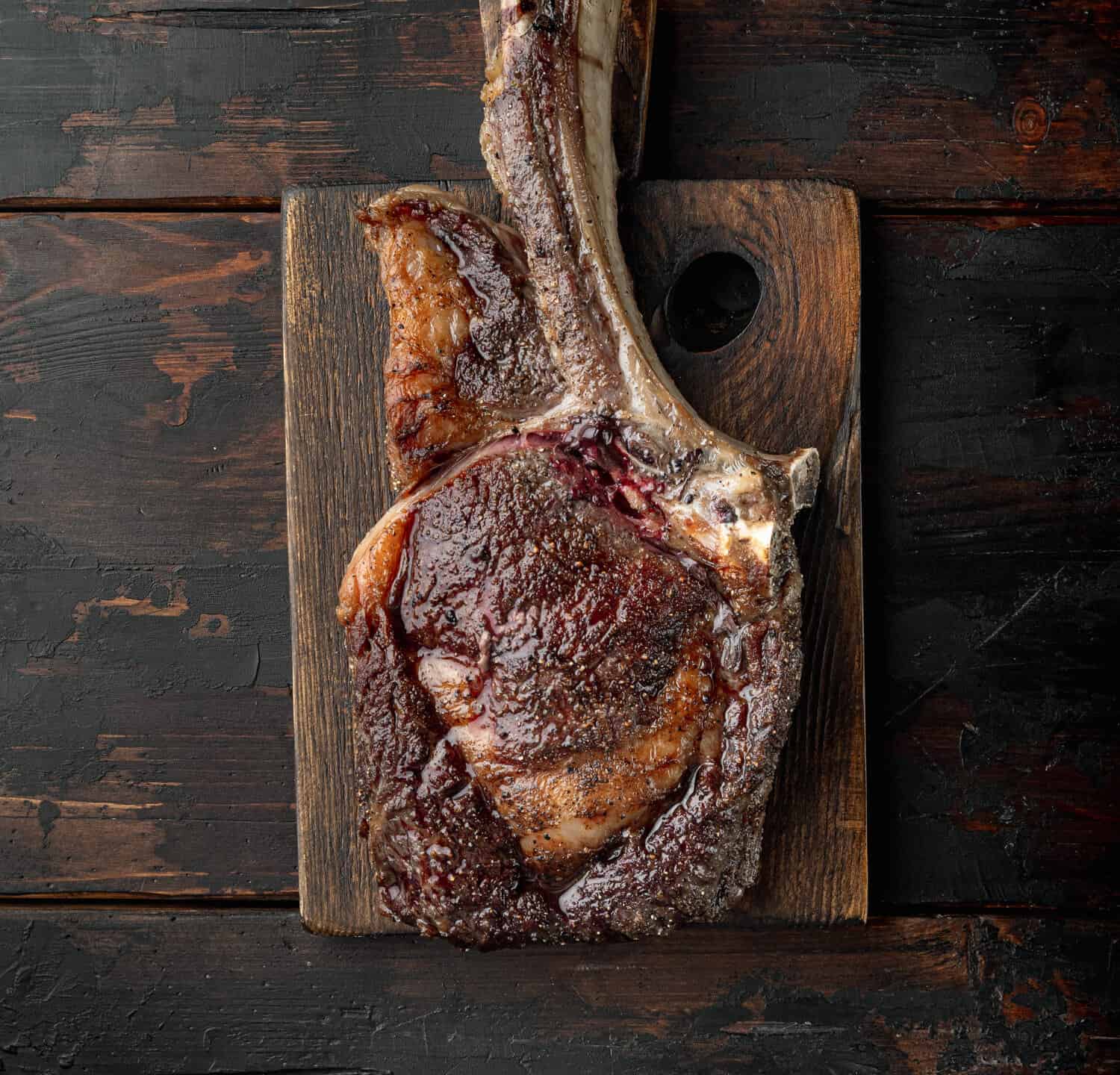 Beautifully seared tomahawk steak, medium rare set, on wooden serving board, on old dark wooden table background, top view flat lay