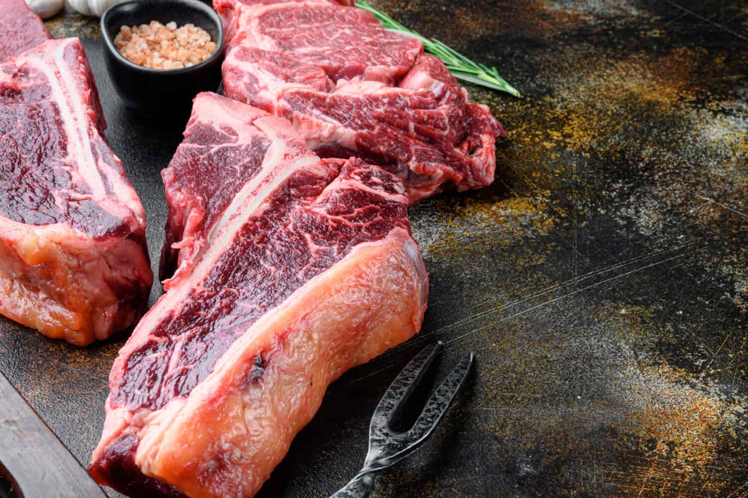 Prime cut tender raw t-bone steak for a BBQ set, on old dark rustic background, with copy space for text
