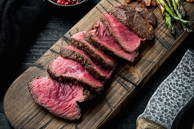 Sliced grilled marbled meat steak Filet Mignon set, with onion and asparagus, on wooden serving board, with meat knife and fork, on black wooden table background