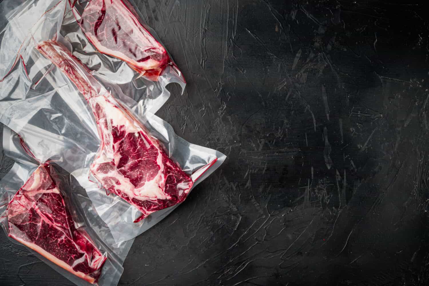 Dry aged steak in a vacuum. Meat products in plastic pack set, tomahawk, t bone and club steak cuts, on black stone background, top view flat lay, with copy space for text