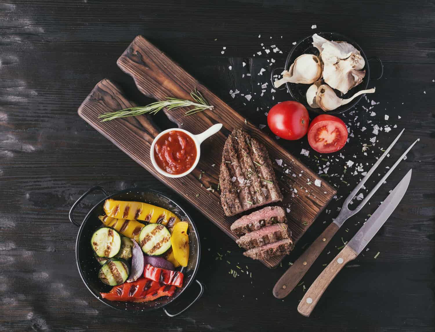 Grilled sirloin steak on wooden serving board with vegetables and spices over rustic wooden background. Top view. 