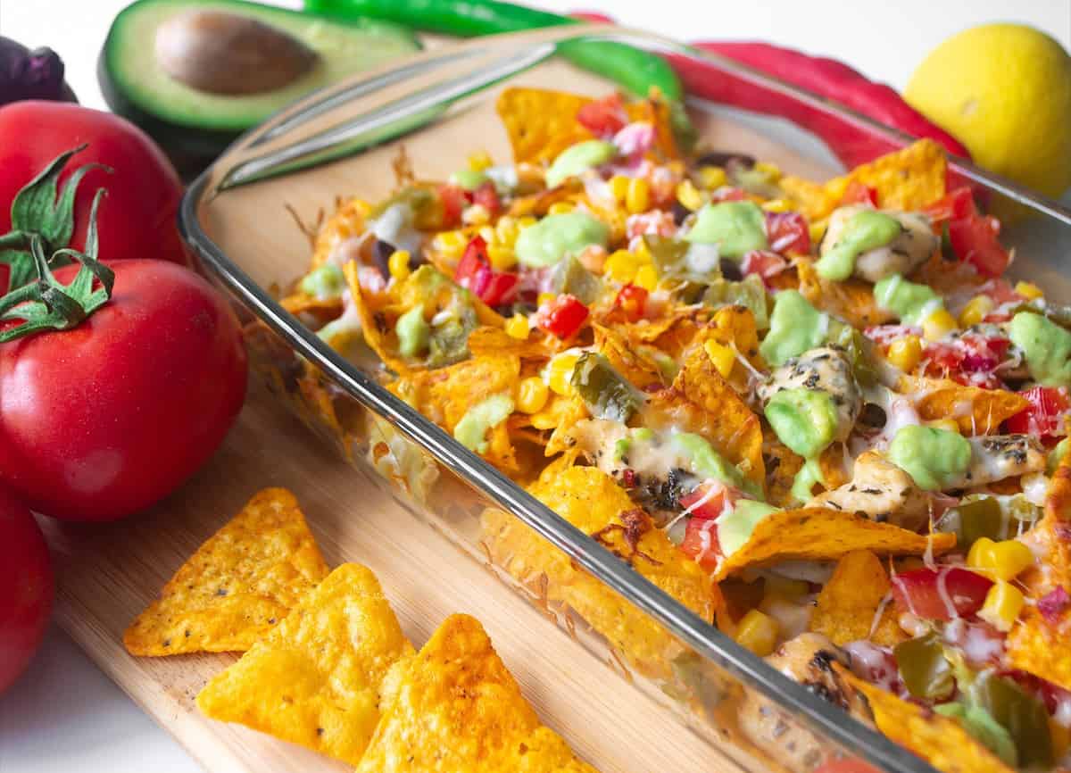 Baked nacho dish in a glass baking pan with corn tortilla chip, pico de gallo salsa, avocado guacamole chipotle, beans, jalapenos and Monterrey jack cheese on a wooden board surrounded by ingredients