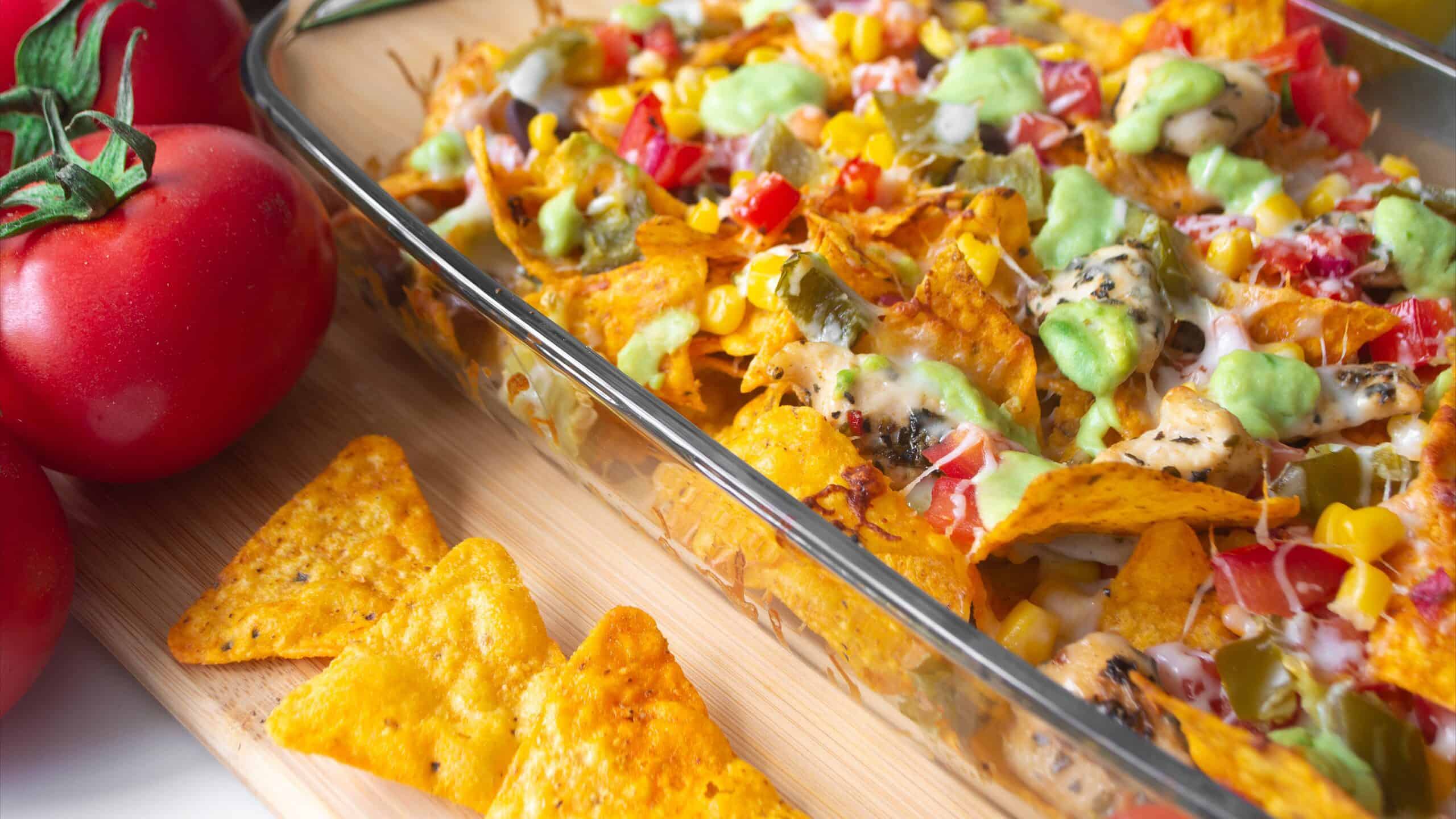 Baked nacho dish in a glass baking pan with corn tortilla chip, pico de gallo salsa, avocado guacamole chipotle, beans, jalapenos and Monterrey jack cheese on a wooden board surrounded by ingredients