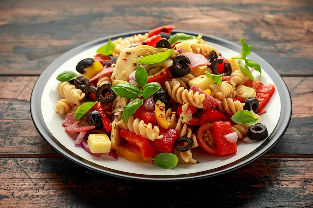Antipasto salad with pasta, tomato, olives, red onion, bell pepper, salami, cheese artichoke and basil on wooden table
