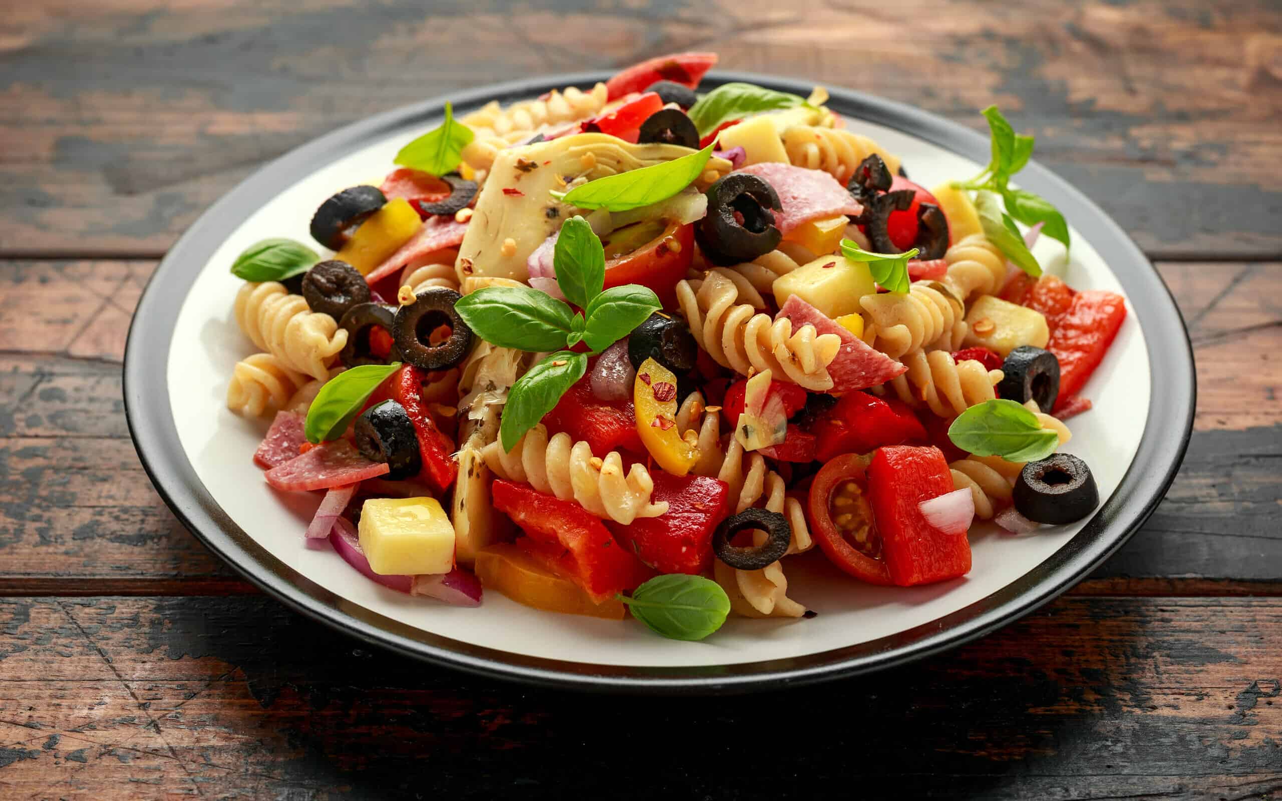 Antipasto salad with pasta, tomato, olives, red onion, bell pepper, salami, cheese artichoke and basil on wooden table