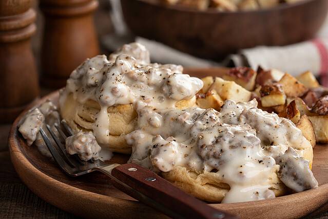 Closeup of biscuits with creamy sausage gravy and fried potatoes on a wooden plate
