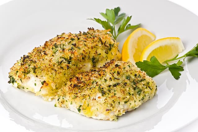 Baked cod with breadcrumbs on a white plate garnished with lemon slices and parsley.