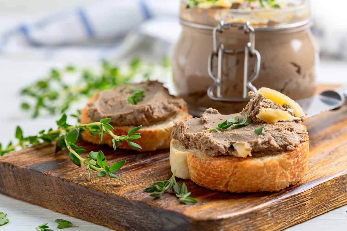 Slices of toasted baguette with turkey pate with thyme and a jar of pate are served on a wooden service board. Concept of delicious homemade food.