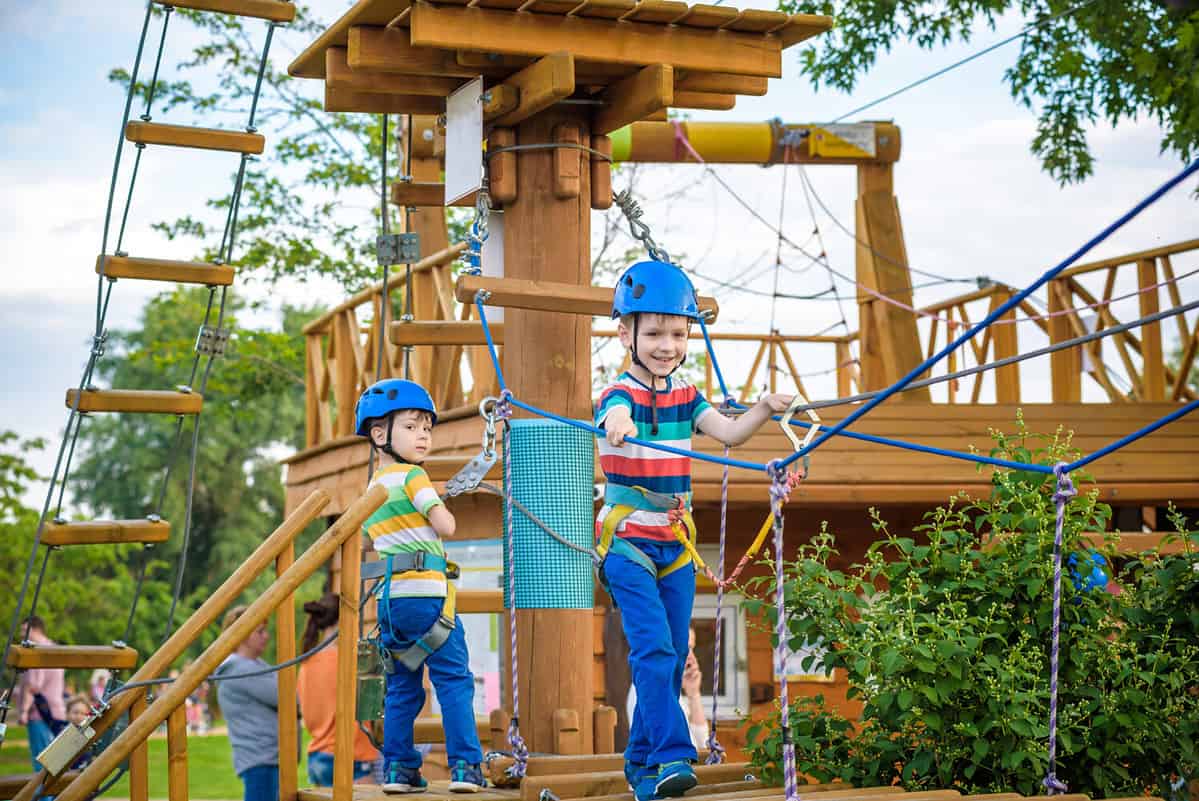 Twin brothers in helmet walks by rope at park sunny summer day in adventure playground. Active healthy leisure time spending concept with children.