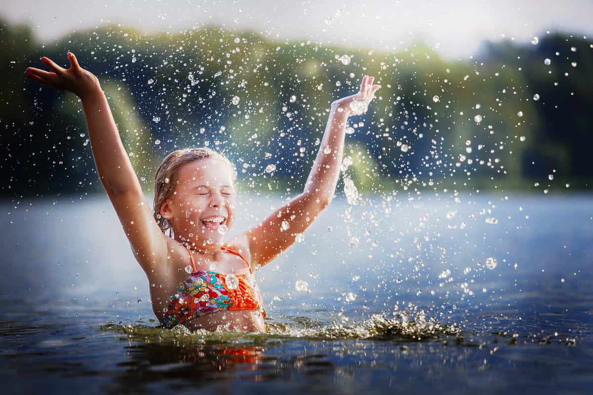little girl smiling playing in the river. A girl with blond hair raises her hands up in the water and splashes water drops.