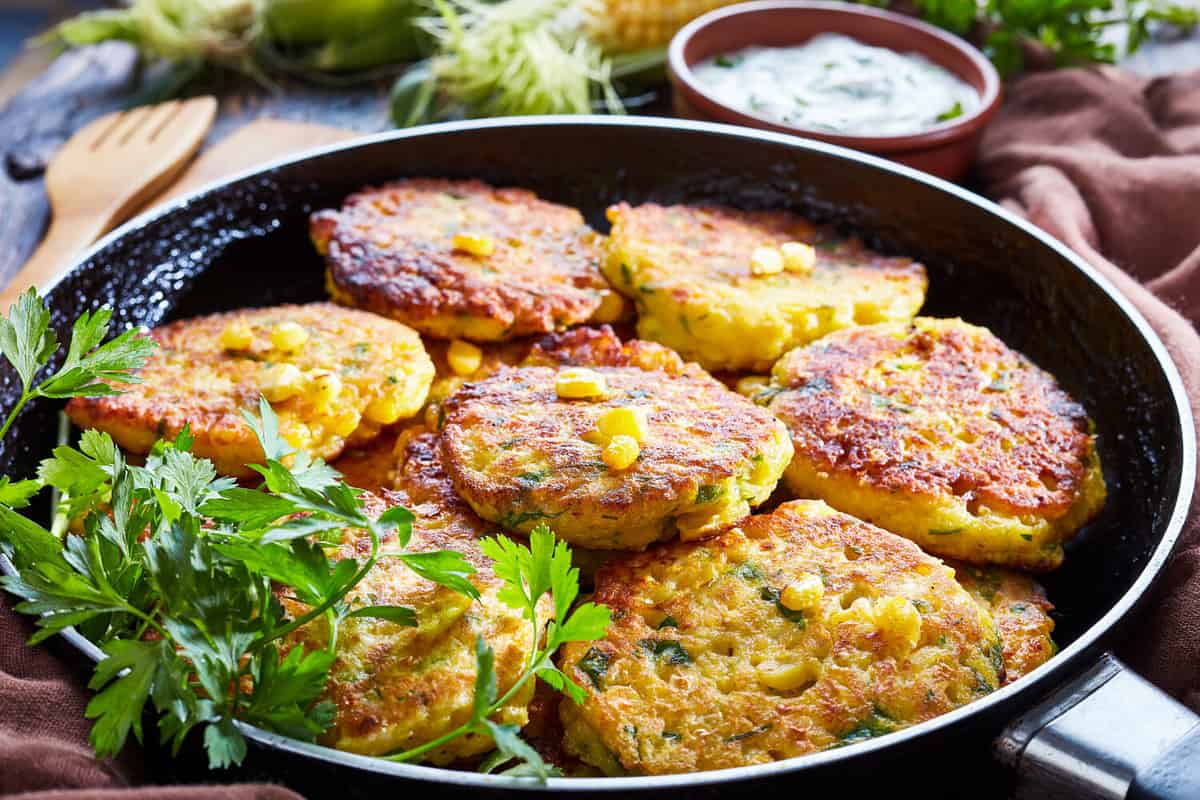 fried corn fritters in a skillet with yogurt dipping sauce and fresh sweet corn in cob on an old wooden kitchen table, close-up