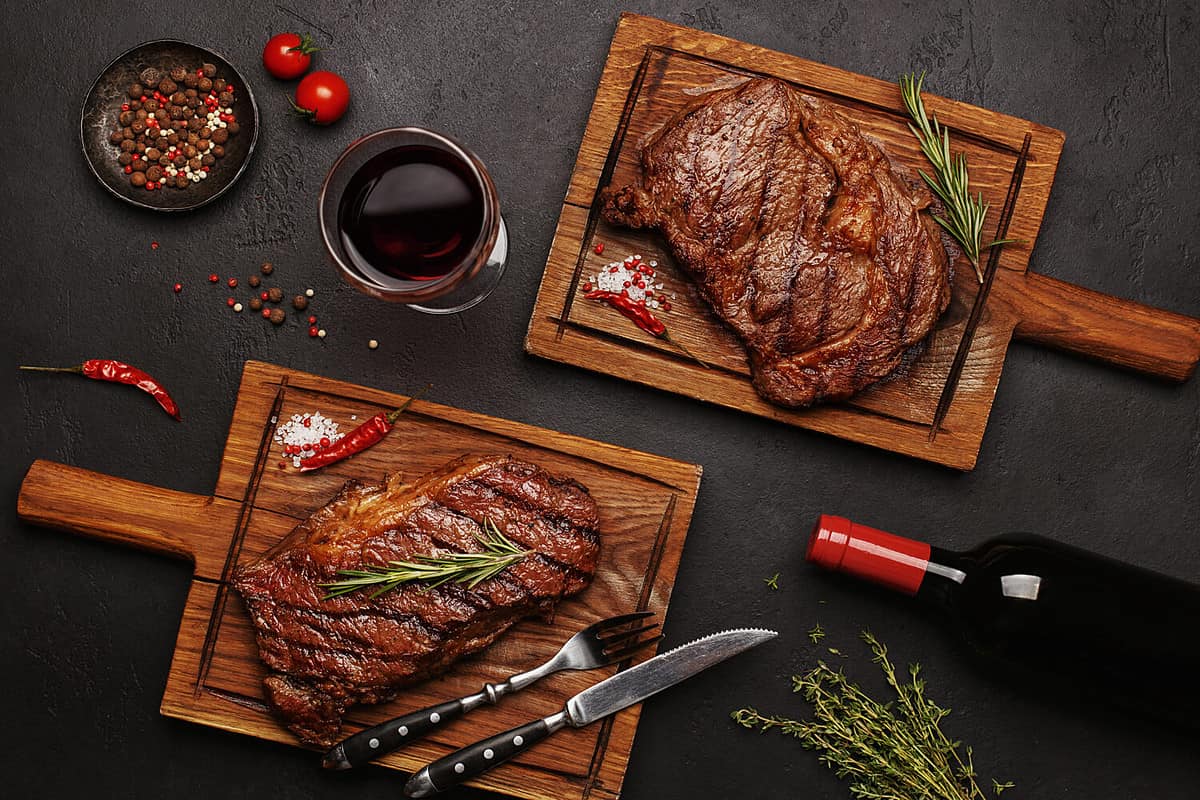 Striploin and Ribeye grilled beef steaks served on wooden boards with glass of wine, bottle of wine, herbs and spices. Top view