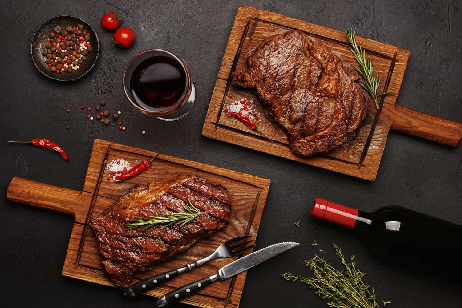 Striploin and Ribeye grilled beef steaks served on wooden boards with glass of wine, bottle of wine, herbs and spices. Top view 