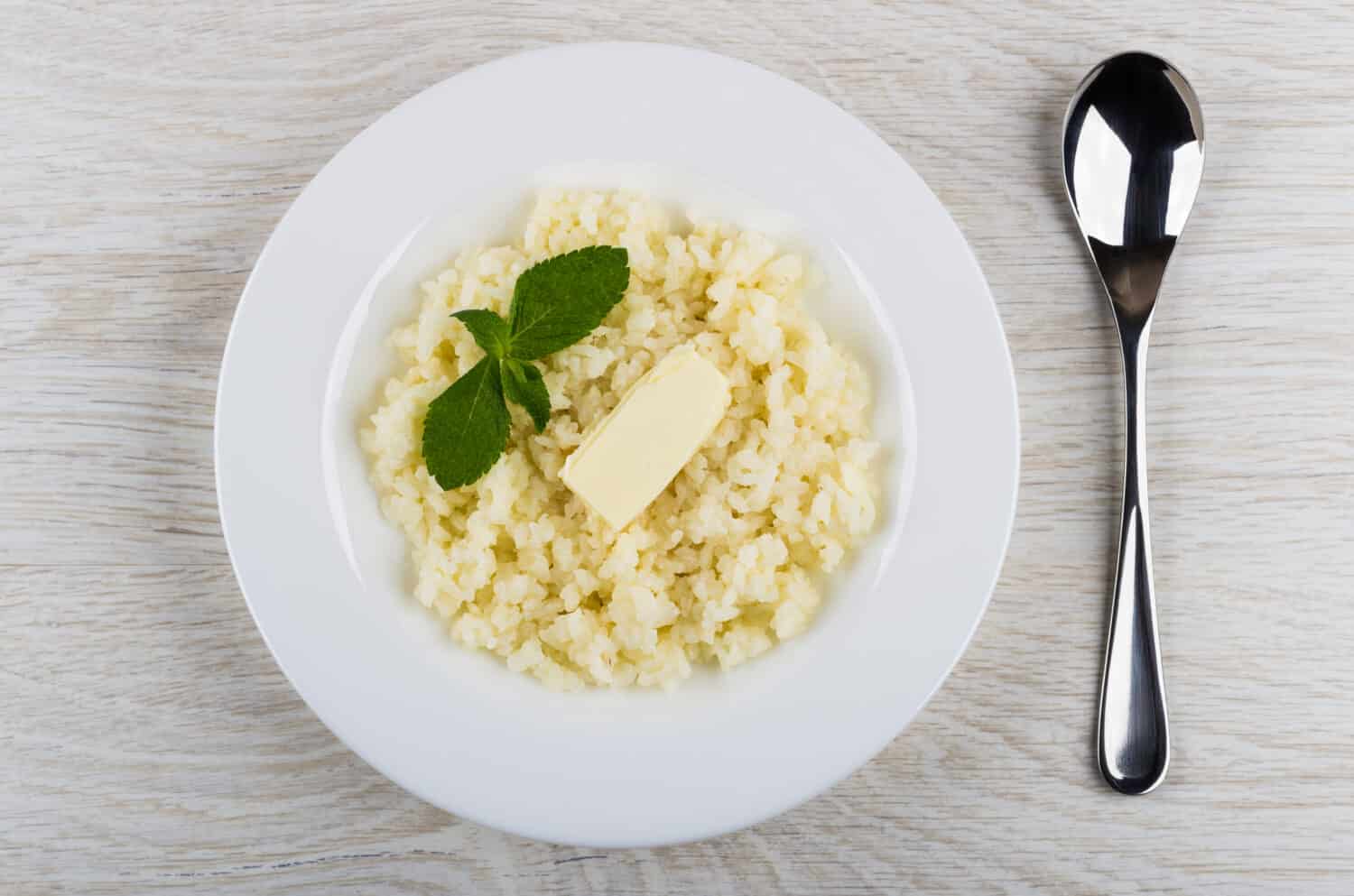 Rice porridge with butter and mint in white plate, spoon on wooden table. Top view