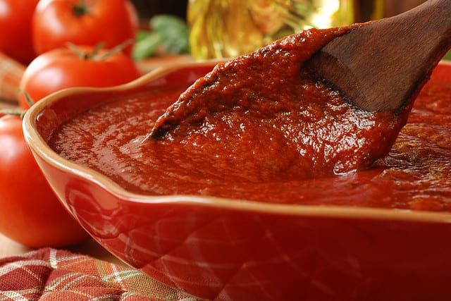 Freshly prepared pasta or pizza sauce in bowl with wooden spoon. Tomatoes, olive oil, and herbs in background. Closeup with shallow dof. Selective focus on sauce in spoon.