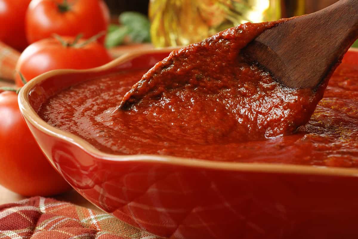 Freshly prepared pasta or pizza sauce in bowl with wooden spoon. Tomatoes, olive oil, and herbs in background. Closeup with shallow dof. Selective focus on sauce in spoon.