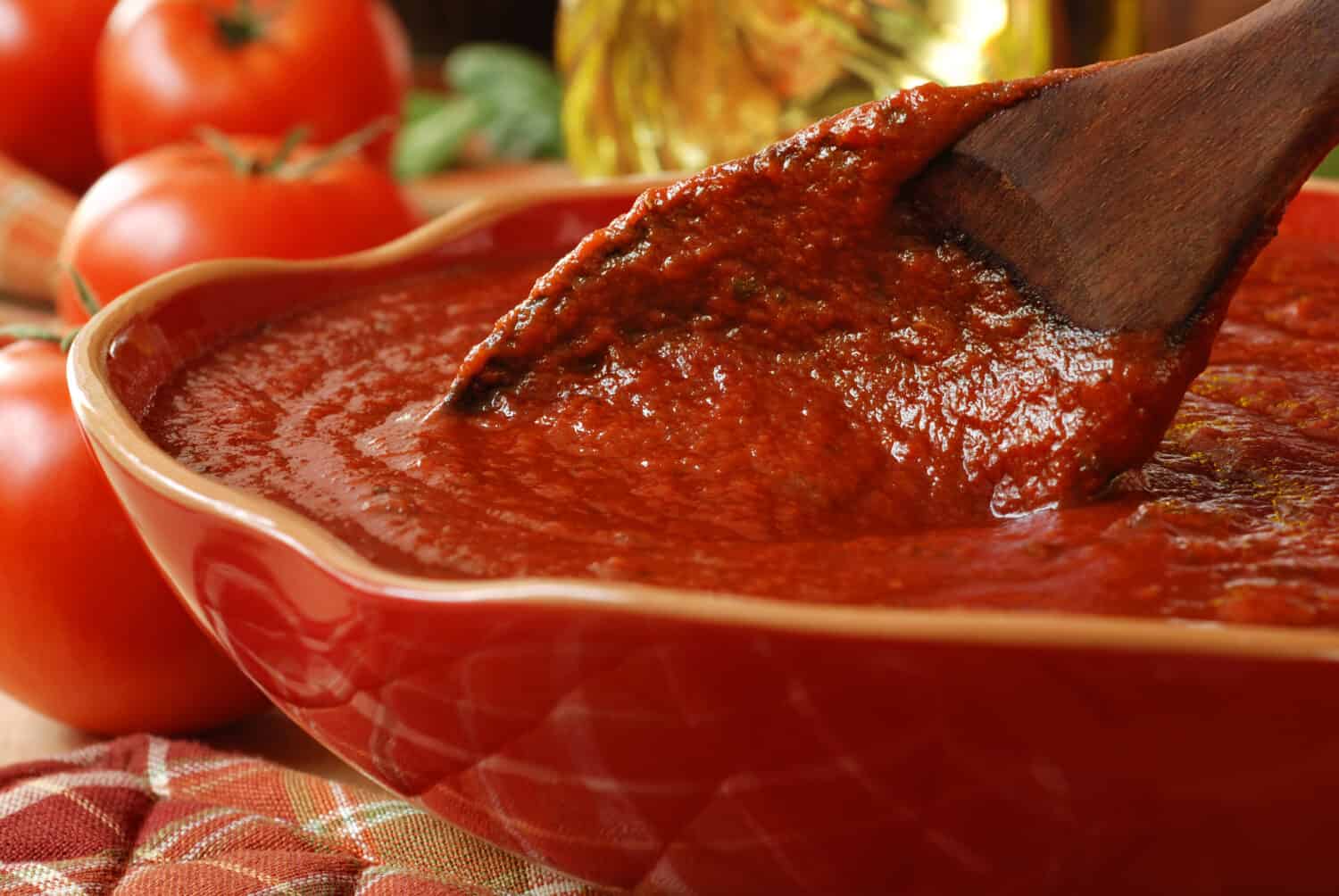 Freshly prepared pasta or pizza sauce in bowl with wooden spoon.  Tomatoes, olive oil, and herbs in background.  Closeup with shallow dof.  Selective focus on sauce in spoon.