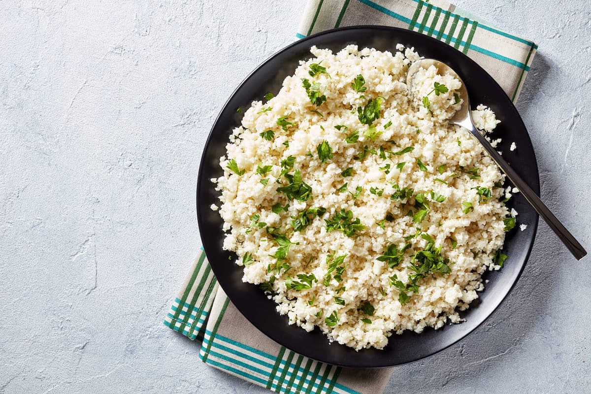 Cauliflower rice or couscous mixed with finely chopped parsley in a black bowl on a white concrete table, healthy eating, low calories food, horizontal view from above, flatlay, empty space