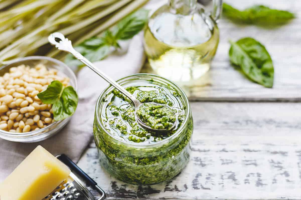 Green fettuccine and Pesto sauce in vintage spoon on glass jar of pesto sauce with ingredients on rustic white wooden table. Traditional Italian pesto recipe for making fettuccine, pasta, bruschetta.